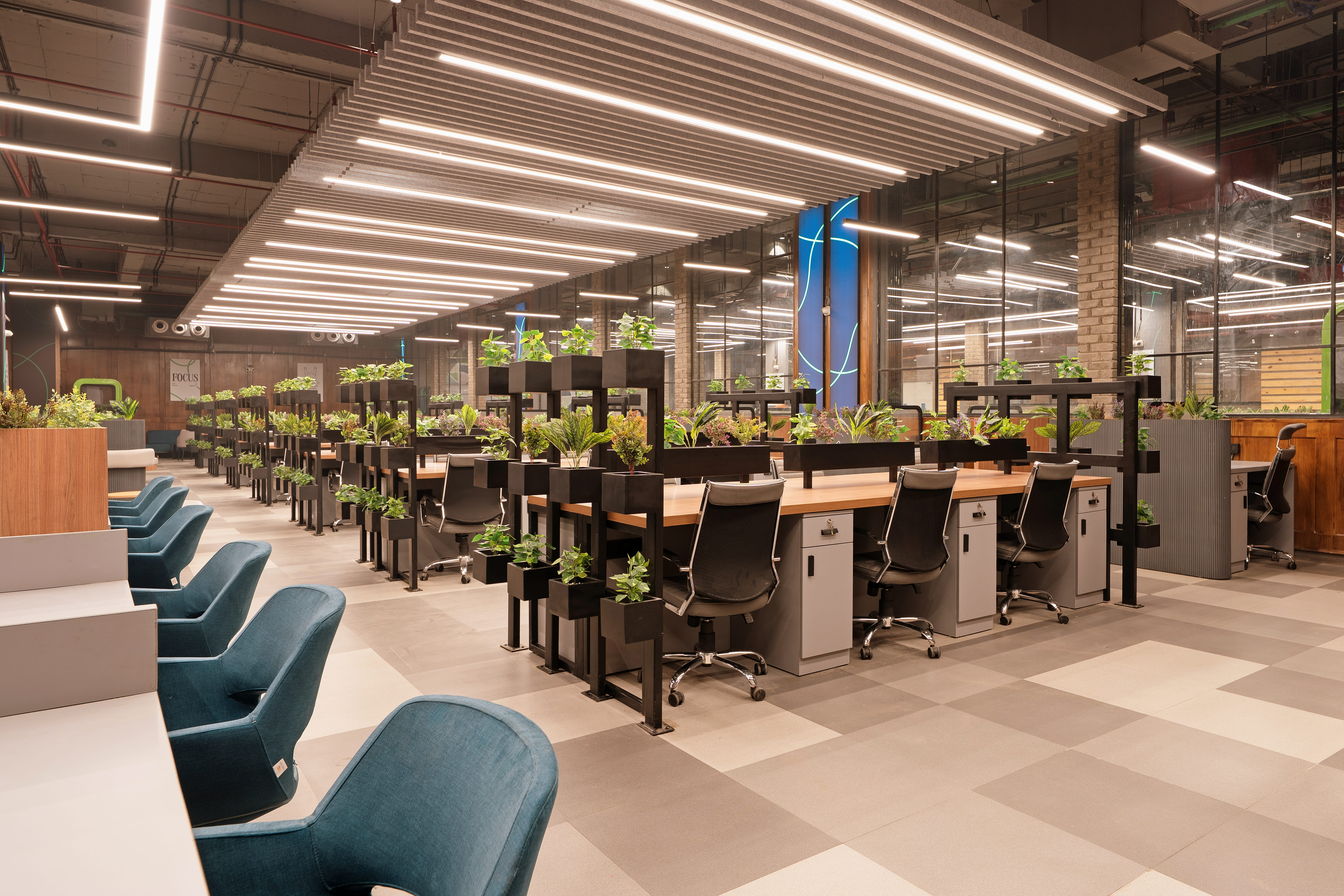 Modern open-plan coworking space with shared desks, office chairs, plants, and bright ceiling lights.